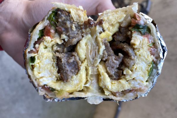 A breakfast burrito cut in half, with sausage, eggs, salsa, and more. A hand is holding the burrito.