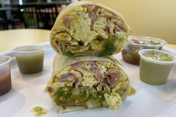 Breakfast burrito stuffed with ham, green peppers, eggs, and more, cut in half and stacked on top of one another, with several salsas in the background