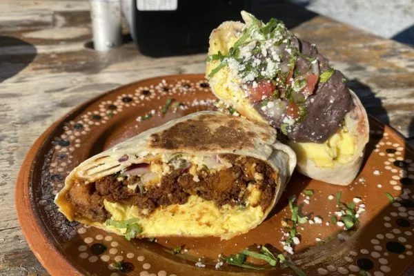 Two breakfast burritos cut in half and stacked, one full of chorizo and egg and the other full of beans and eggs. The burritos are on a brown patterned plate on a wooden table.