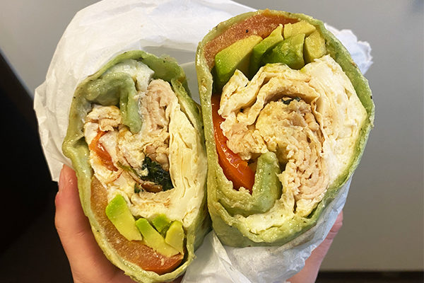 Breakfast burrito in a green wrap sliced in half and displayed to the camera, full of deli meat, avocado, tomato, and more