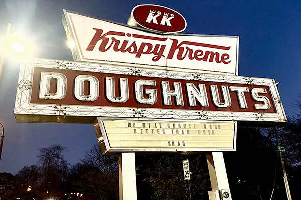 A Krispy Kreme sign against a dusk sky, reading "We will bounce back better than ever - Shaq"