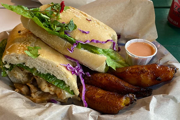 Chicken cubano sandwich with fried plantains and a white cup of pink sauce