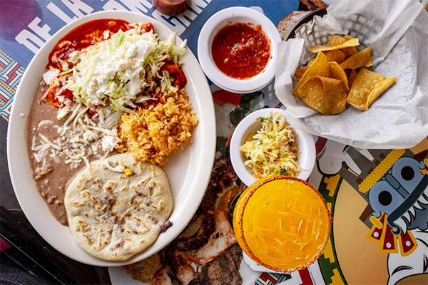 Plate of pupusa, rice, and beans next to a platter of chips and salsa and a cocktail