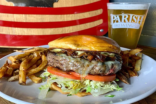 A cheeseburger with caramelized onions, tomato, cheese, lettuce, and tomato with fries and a glass of beer behind it