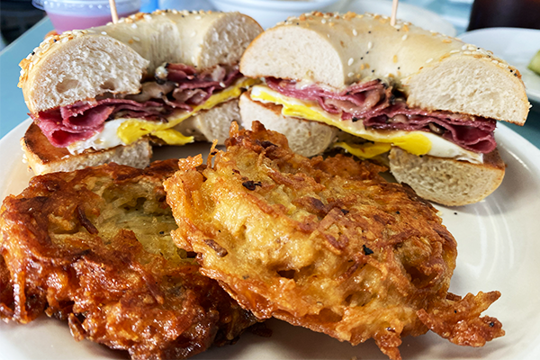 A bagel sandwich cut in half and topped with pastrami, egg, and cheese with golden potato pancakes in the foreground