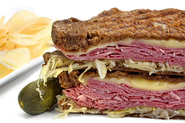 A pressed sandwich on rye bread, topped with corned beef, sauerkraut, and swiss cheese. There's a pickle and potato chips in the background