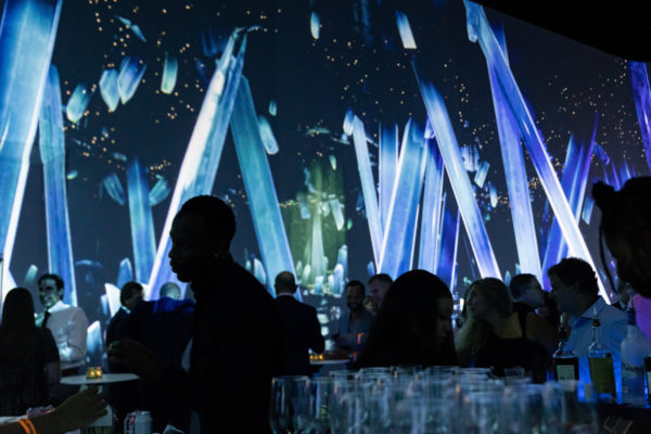 People sitting at dinner tables with glasses in the foreground and a projected screen in the background