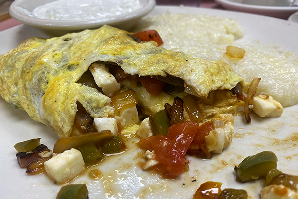 Onions, peppers, tomatoes, feta, and gyro meat in an omelet with tzatziki sauce and grits in the background