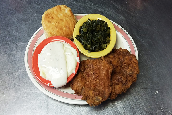 Fried pork chop on plate with greens, mashed potatoes with gravy, and a biscuit