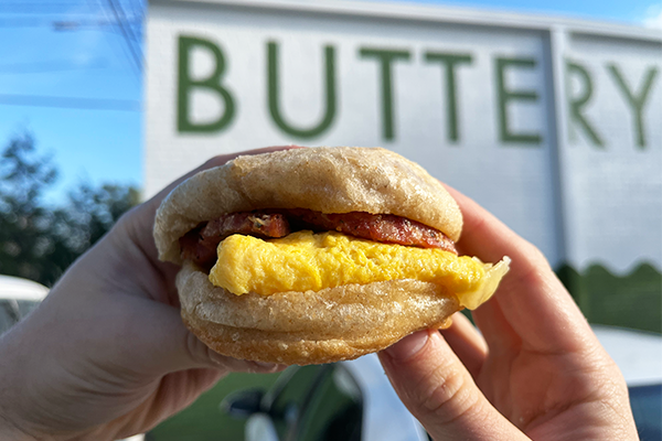 Breakfast sandwich on english muffin being held up in front of sign reading "Buttery"