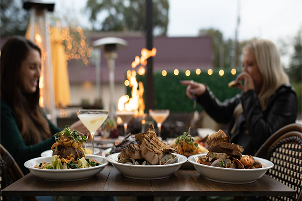 Several dishes on table with two drinks while two women are talking and a firepit is in the background
