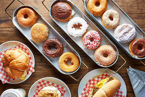 Several trays of various donuts, with plates with breakfast sandwiches on below them