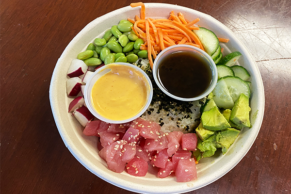 Poke bowl with tuna, avocado, cucumber, carrots, edamame, radish, spicy mayo, and house marinade on a brown table