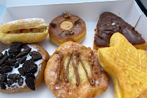 Six donuts in a box. Clockwise from top left: strawberries and cream in a donut, peanut butter cup donut, A-shaped chocolate cream-filled donut, an orange star, a fritter with apple in the middle and a caramel drizzle, and a cookies and cream donut with pieces of dark Oreo cookie