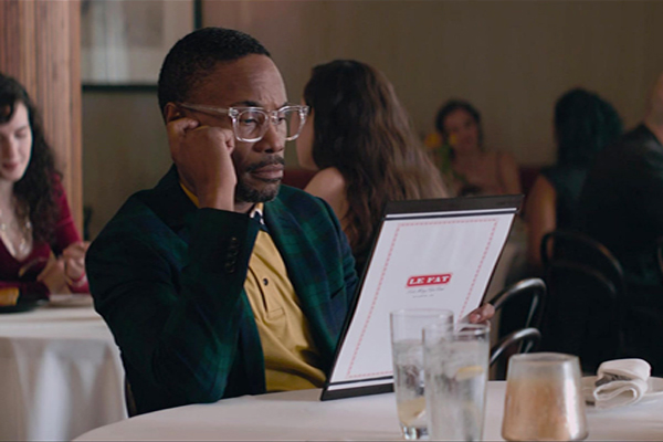 A man in glasses sitting at a table in a restaurant reading a menu that says "Le Fat"