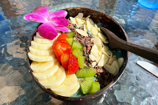 Blue yogurt bowl topped with bananas, strawberries, kiwis, cacao nibs, and slivered almonds with a pink flower