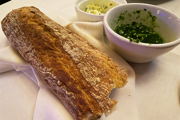 Loaf of bread on a white napkin next to a white cup of green pesto and a smaller ramekin of butter with green herbs in it