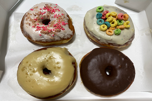 Four donuts on white paper: one with red powder, another with rainbow cereal, one with caramel frosting, and another with chocolate frosting