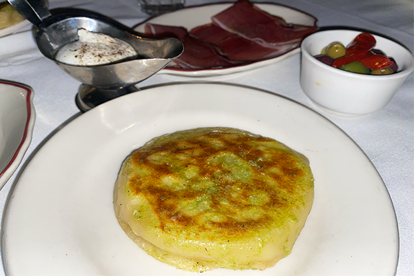 A circular piece of pressed focaccia on a plate with ricotta, speck, and olives/peppers in the background