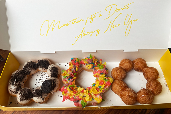 Three uniquely-shaped donuts in a yellow box, one topped with white glaze and black cookies crumbs, the next topped with rainbow cereal, and the third topped with a caramel glaze
