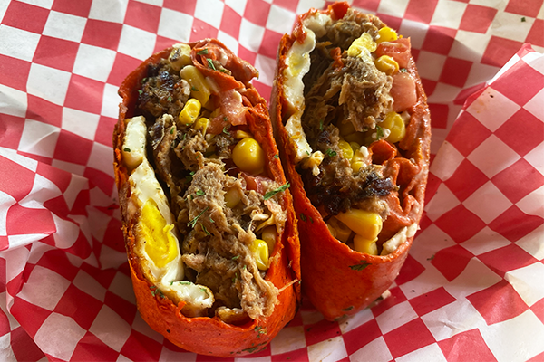 Breakfast burrito with a red wrap, split in half and full of pulled pork, corn, tomatoes, feta, and green garnish