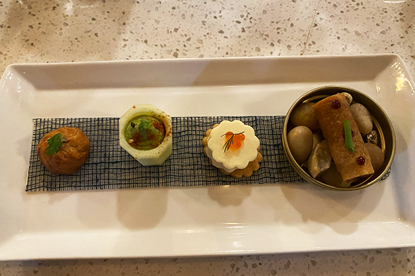 An assortment of four bite-sized apps (mushroom pastry, cucumber filled with avocado, a savory cracker topped with a mousse, and a meat-filled eggroll on rocks)