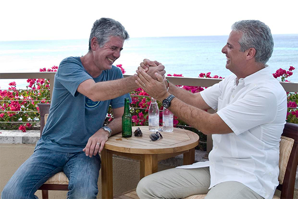 Anthony Bourdain and Eric Ripert holding hands while sitting at a table overlooking a body of water