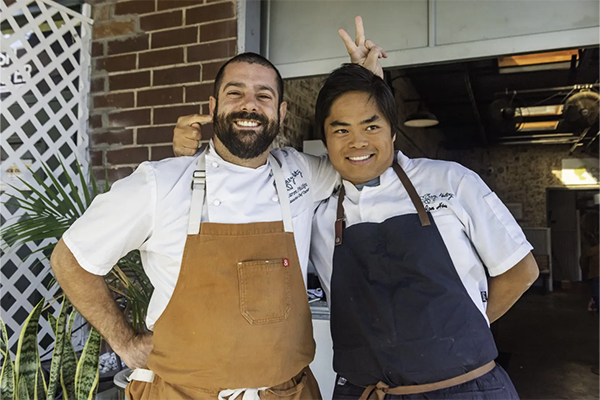 Two chefs with their arms around each other, one giving the other bunny ears and the other sticking his finger in the other's ear