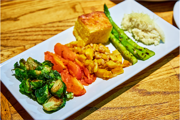 A rectangular dish with Brussels sprouts, carrots, asparagus, and more with a piece of cornbread