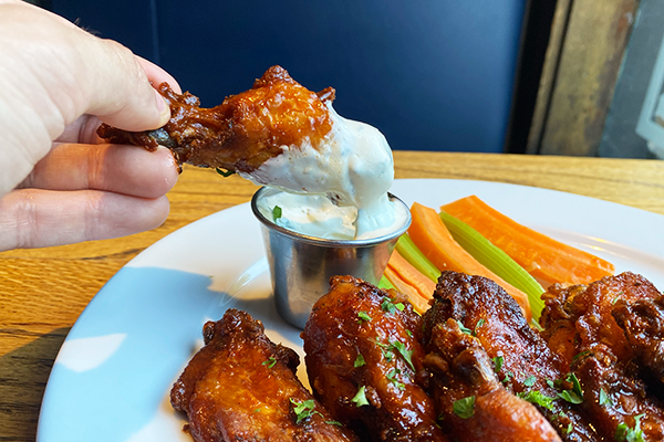 A wing being dipped into ranch dressing with wings in the foreground and carrots/celery in the background