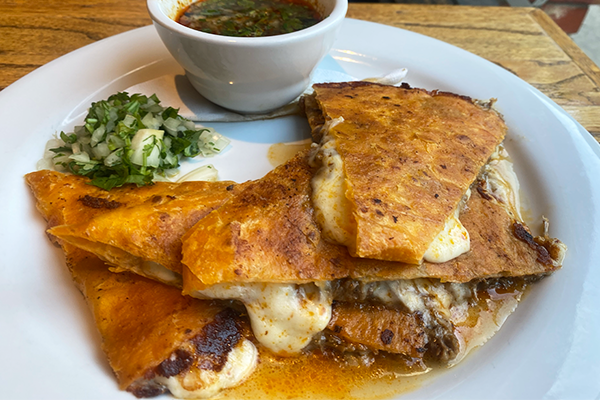 A birria quesadilla sitting on a plate with cilantro and onions on the side and a bowl of beef broth in the background