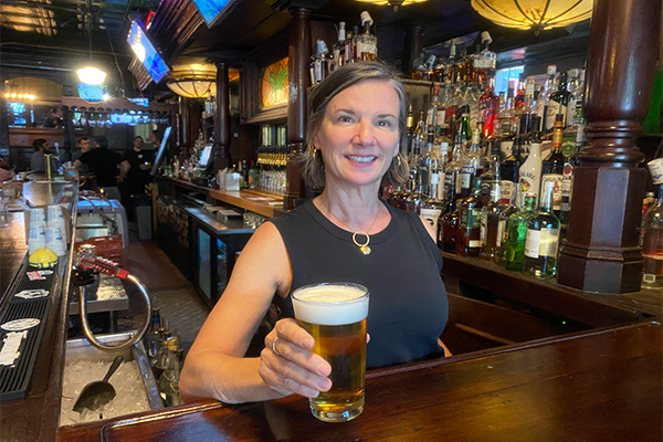 A woman in a black shirt standing at a brown bar holding a glass of beer