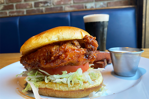 A fried chicken sandwich with lettuce and tomato on a white plate with sweet potato fries and a Guinness in the background