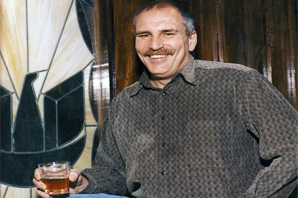 A man holding a glass with a brown liquid smiling at a camera
