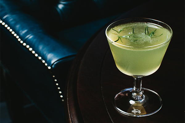 Green cocktail with a sprig of rosemary floating in it served atop a table in front of a black leather chair