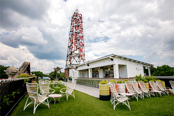 Rooftop bar with turf on the ground, several white chairs, and a white building with a tall structure reading "Hotel Clermont" in red letters 
