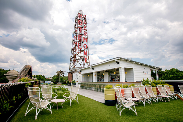 Rooftop bar with turf on the ground, several white chairs, and a white building with a tall structure reading "Hotel Clermont" in red letters