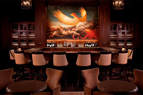 Dimly-lit bar with a colorful mural with a flying bird over the back. There are several chairs lining the bar and bottles arranged on top of it