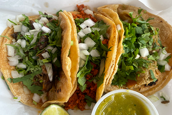 Three tacos with different meats and topped with cilantro and onions and served with a side of green salsa and a lime wedge